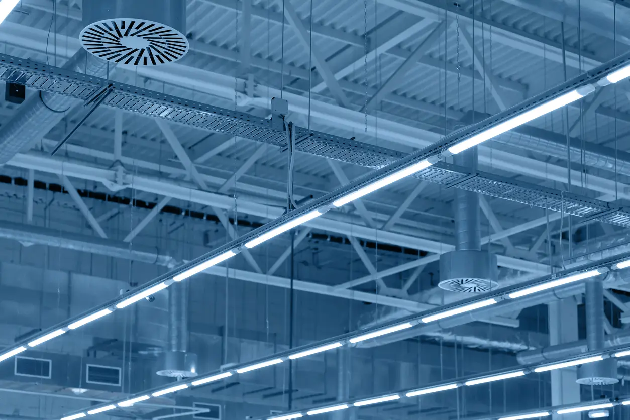 Imagem de luminárias lineares de LED instaladas no teto de um ambiente industrial, com estrutura metálica aparente e iluminação branca intensa.