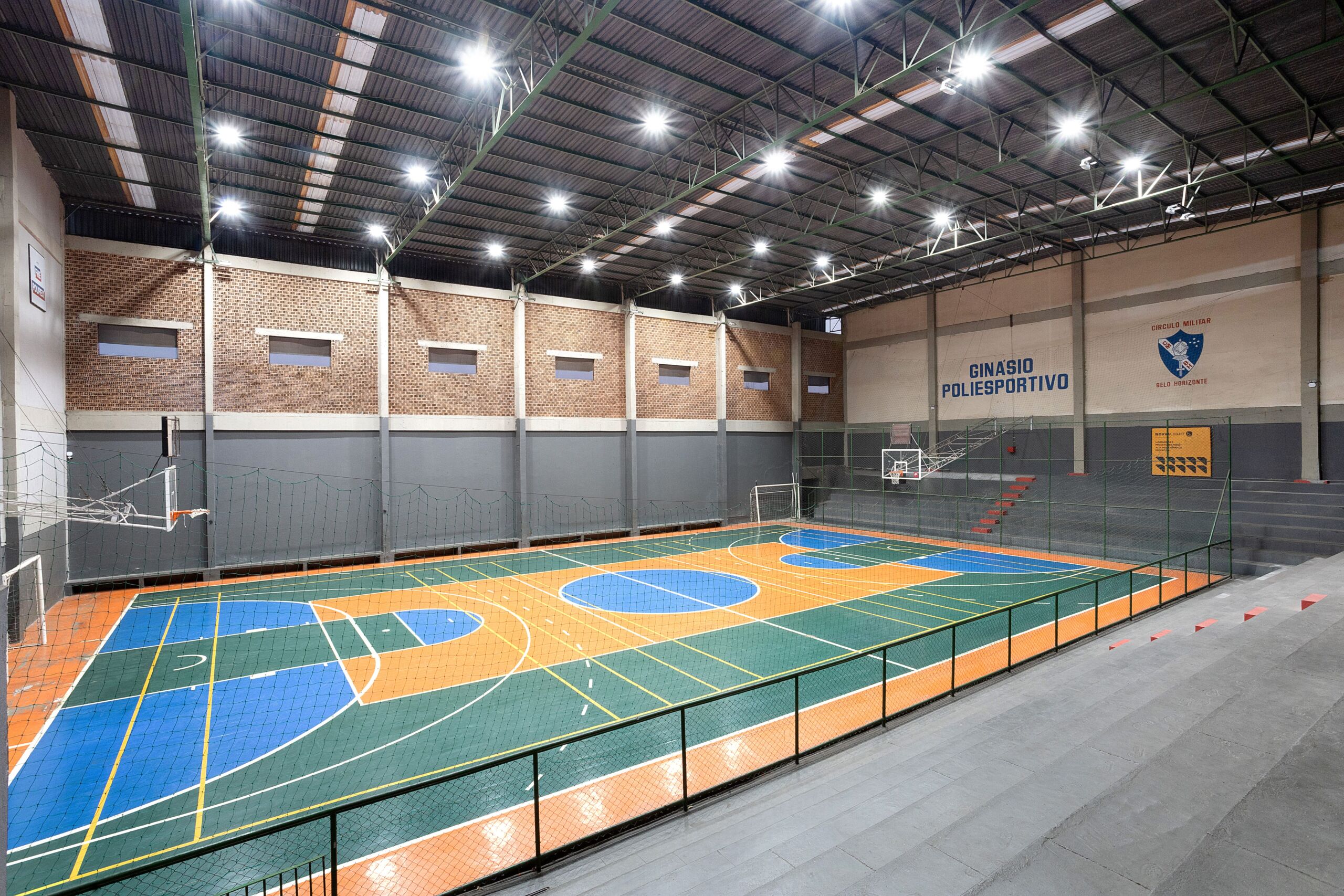 Imagem de um ginásio poliesportivo coberto, com a quadra demarcada para diversos esportes e iluminação forte instalada no teto.