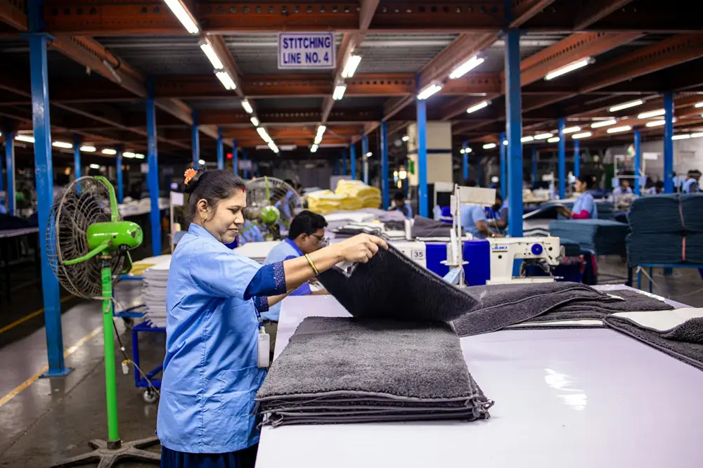 Mulher com uniforme azul em uma indústria têxtil manuseando tecidos que estão em cima de uma mesa.