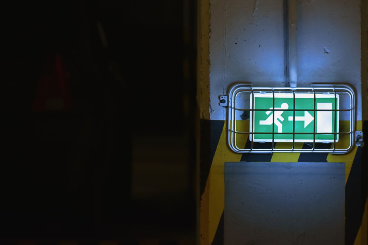 A imagem mostra uma placa luminosa de sinalização de saída de emergência, instalada em um ambiente escuro.