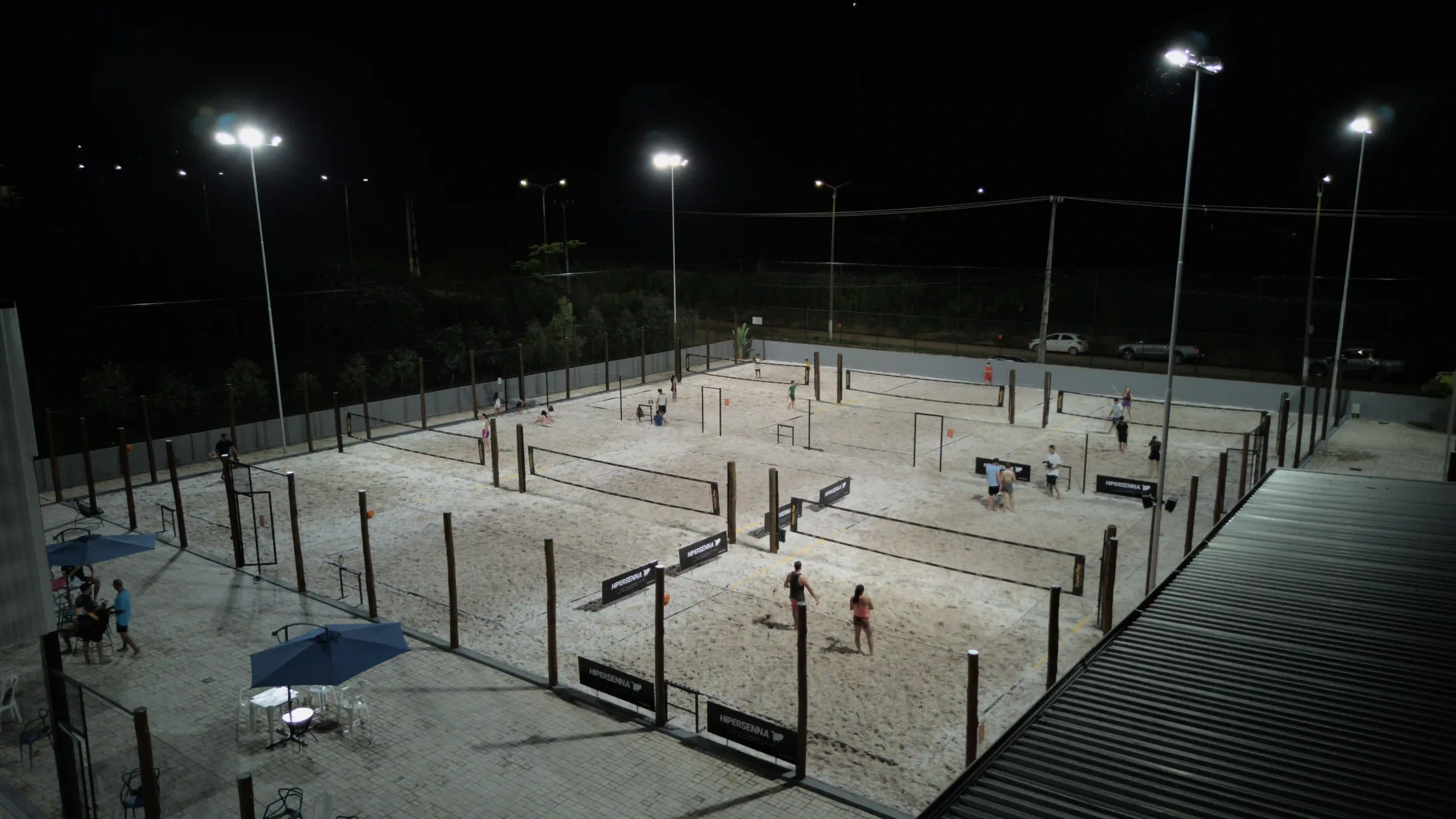Imagem de uma quadra esportiva à noite, bem iluminada, com pessoas praticando esportes.
