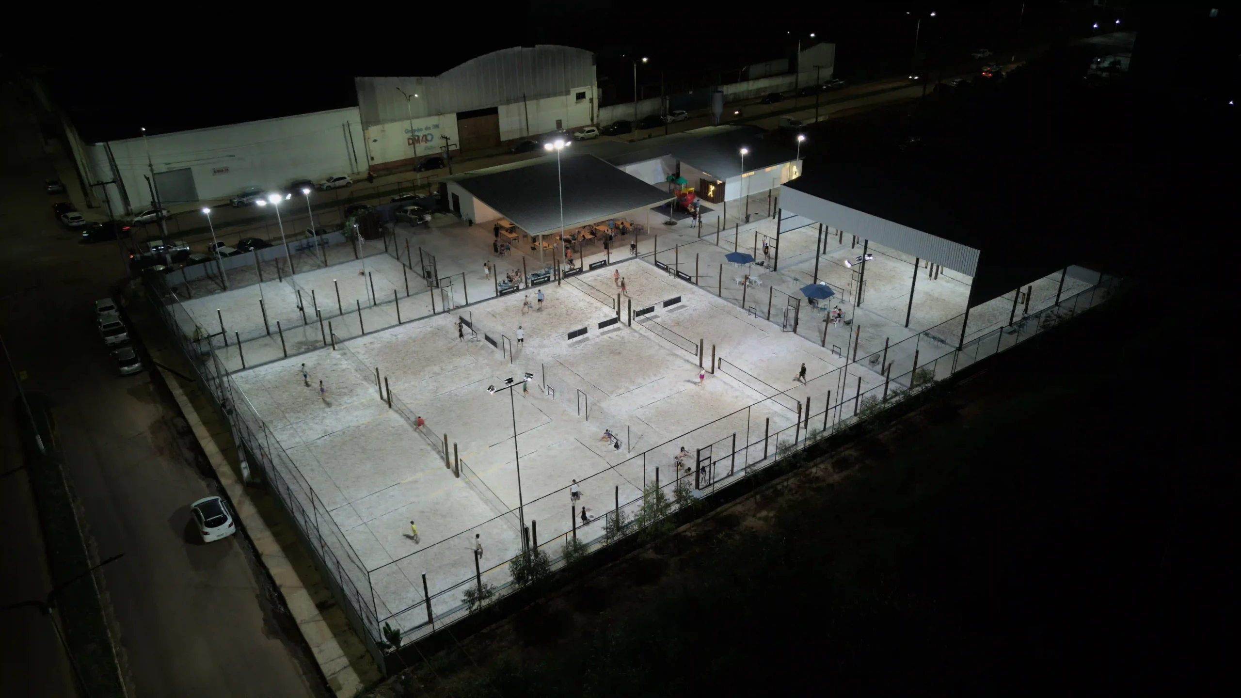 Imagem de uma quadra esportiva à noite, bem iluminada, com pessoas praticando esportes.