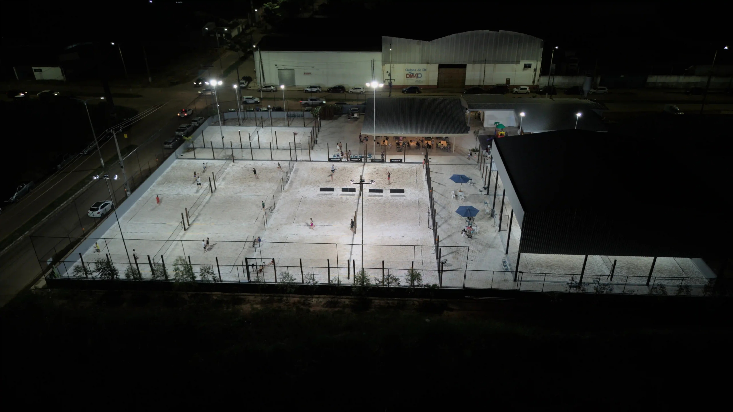 Imagem de uma quadra esportiva à noite, bem iluminada, com pessoas praticando esportes.